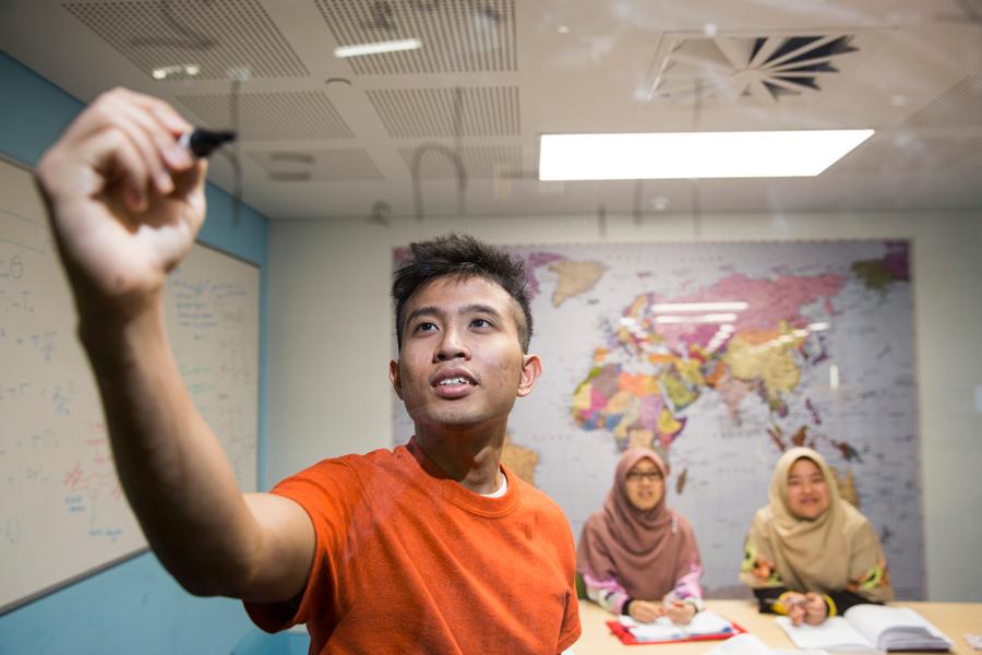 Student writing on a glass board with two students in the background, behind them a map of the world