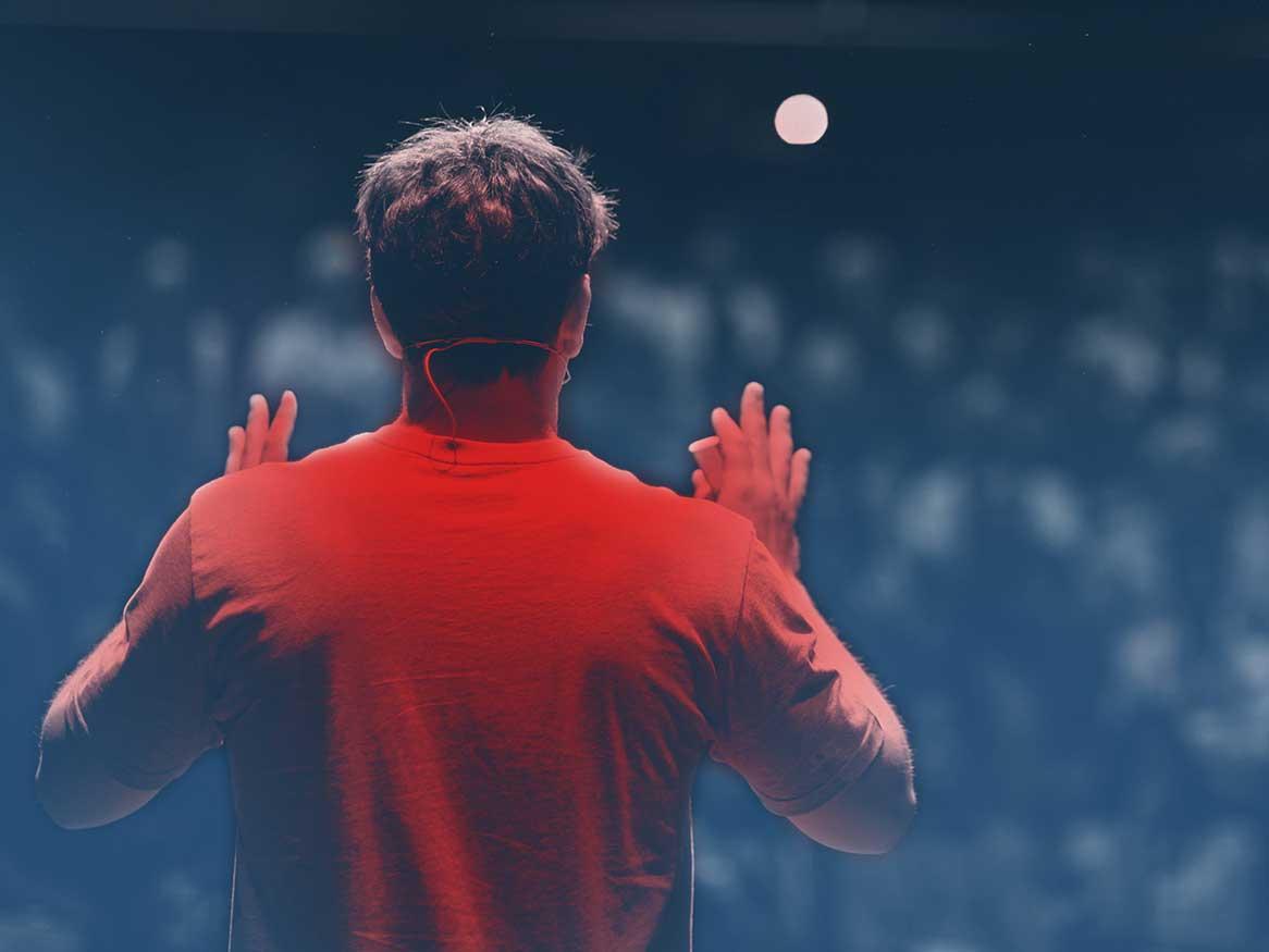 3MT promo image - a man in a red tshirt is shown from behind speaking to a fuzzy large audience