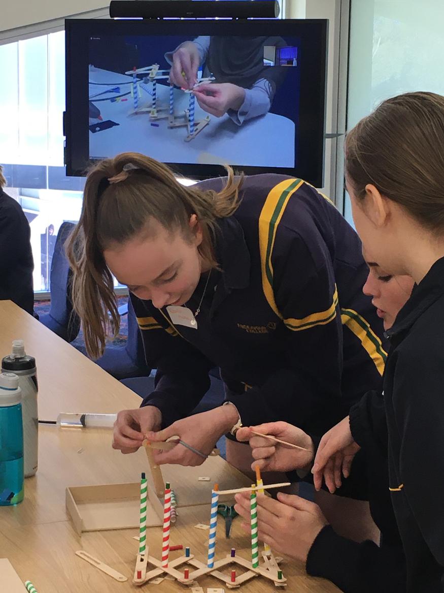 Two female high school students working on a construction project with a tv screen in the background livestreaming the instructor