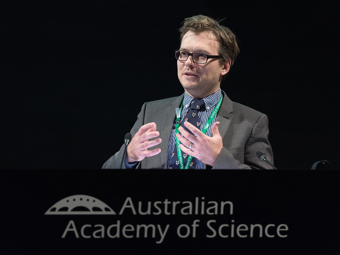 Joshua Ross wearing a suit speaking on part of a panel, with the text Australian Academy of Science