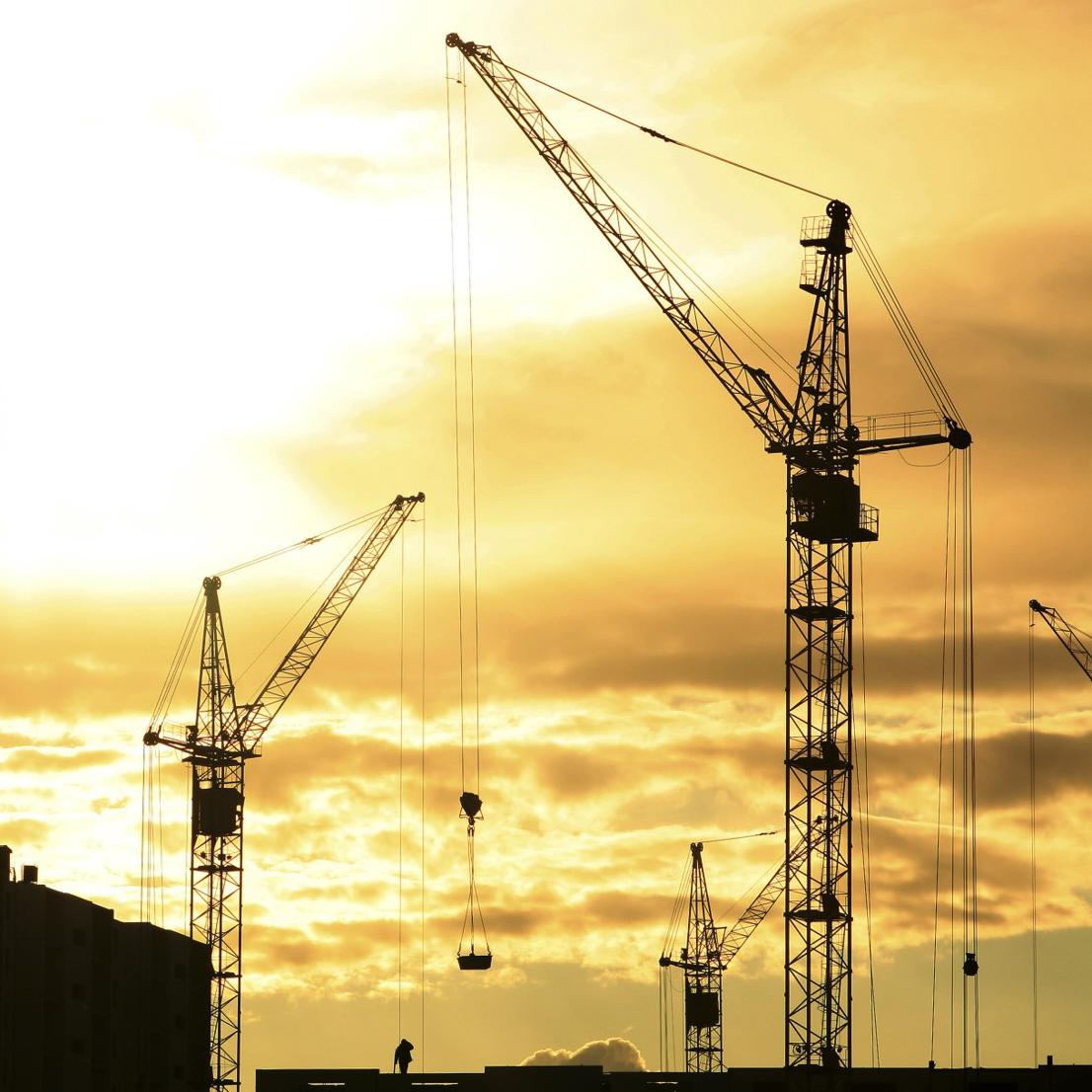 Picture shows a number of construction cranes against a yellow sunset cloudy sky