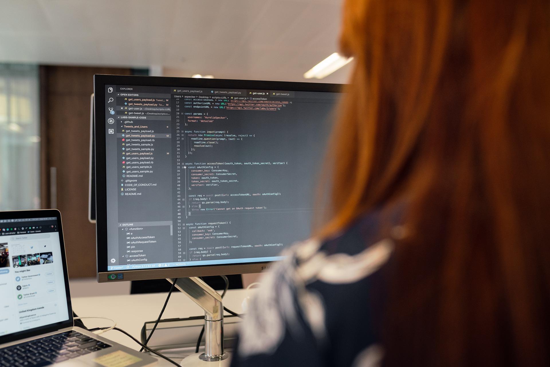 Woman with red hair seen from behind as she sits in front of a computer, coding