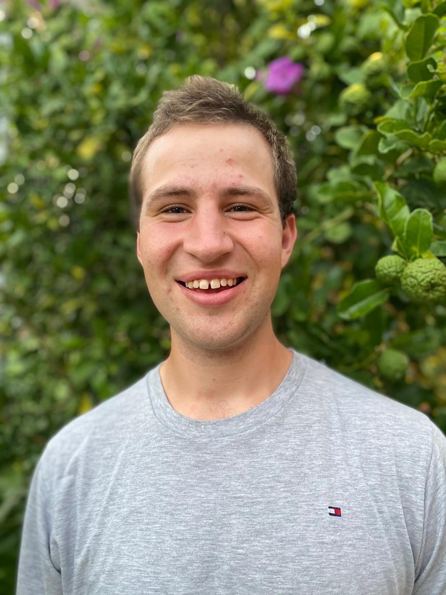 photo of xavier barnett. he is wearing a grey tshirt and is smiling. there are bushes in the background and a purple flower