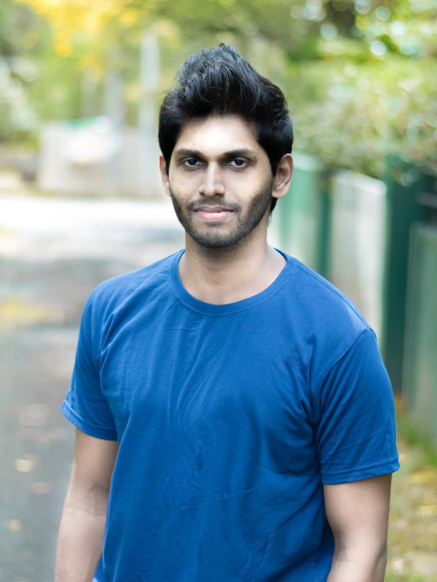 a man with black hair is wearing a blue tshirt and he is standing outside. it is autumn.