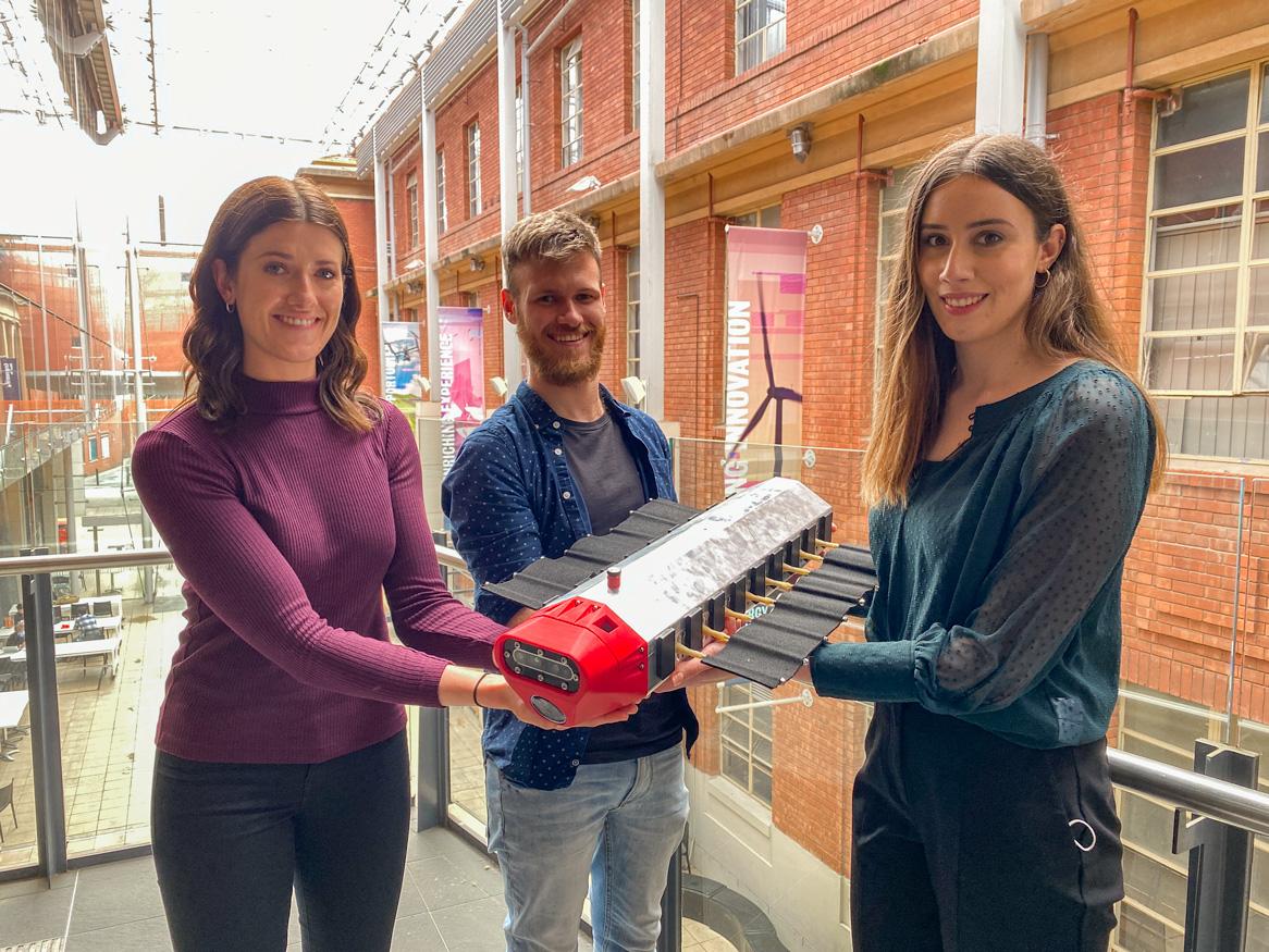 two women and one man are holding a prototype rectangular mechanical object. they are smiling