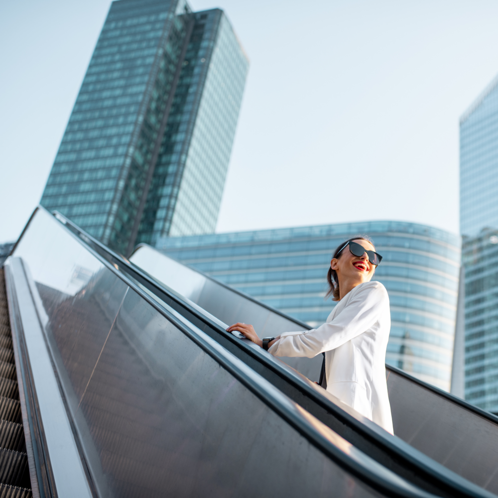 Lady in white jacket, wearing black sunglasses, riding up outside city escalator