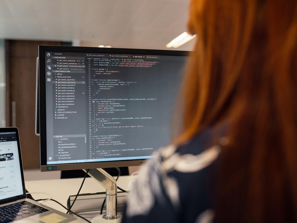 Woman with red hair seen from behind as she sits in front of a computer, coding