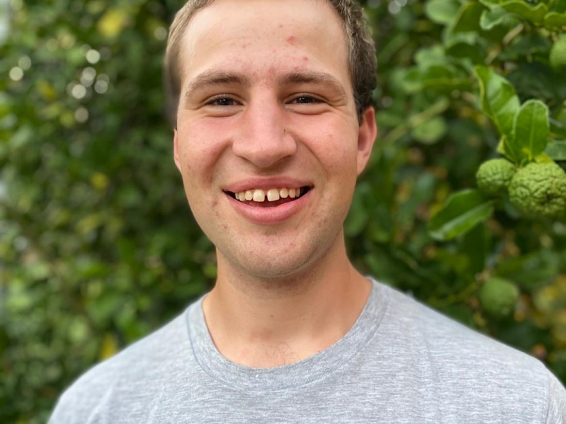 man smiling wearing a grey tshirt standing in front of a green bush